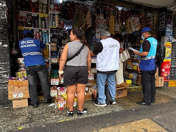 Personal con chalecos de fiscalización y sanidad inspecciona un puesto callejero de comestibles y golosinas frente a dos personas