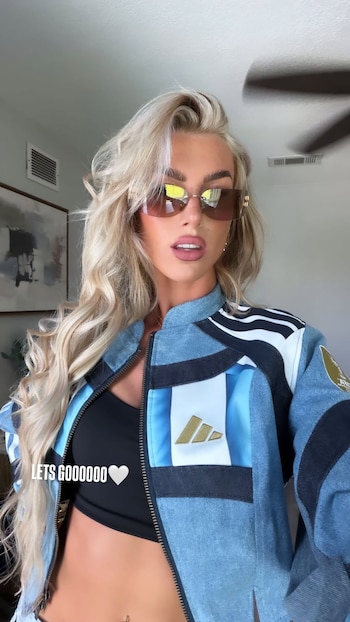 Mujer rubia con gafas de sol, chaqueta deportiva azul y blanca, top negro y ombligo descubierto con piercing. Fondo con pared y cuadro
