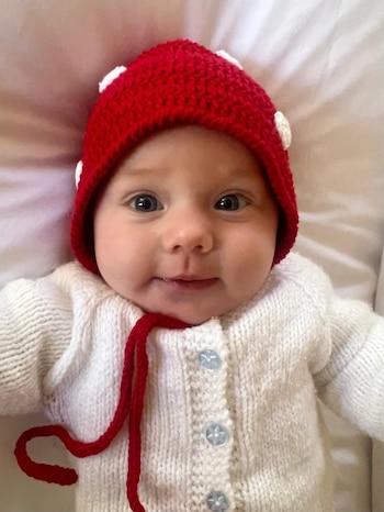 Primer plano de un bebé sonriente con ojos azules, un gorro de lana rojo con puntos blancos y un cárdigan blanco tejido