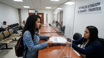 Una mujer joven con cabello rizado y chaqueta de mezclilla entrega documentos etiquetados "ACCION DE TUTELA SOLICITUD" a una empleada tras un mostrador con divisor.