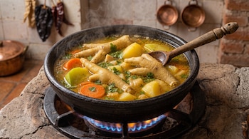 Olla de barro sobre una hornilla encendida con caldo, patas de pollo, papas, zanahorias, chayote y una cuchara de madera.