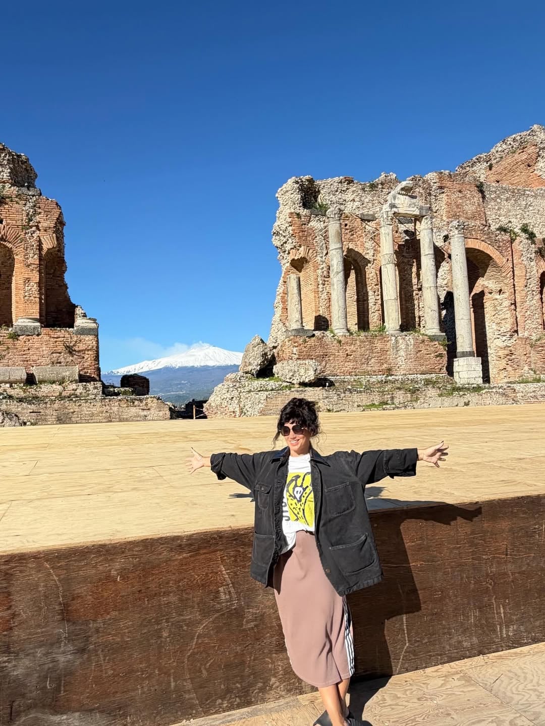 Siciliani posó sonriente con los brazos abiertos frente a las ruinas de un teatro antiguo en Sicilia, con el majestuoso volcán Etna nevado al fondo