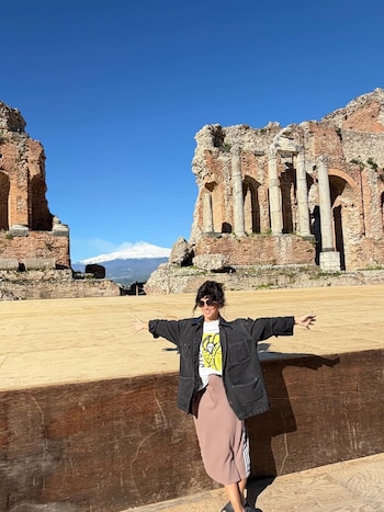 Griselda Siciliani sorrindo com os braços estendidos diante das ruínas de um antigo teatro de pedra. Ao fundo você pode ver o Monte Etna coberto de neve