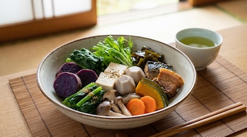 Un plato hondo con boniato morado, tofu, vegetales verdes, zanahorias, calabaza, setas, algas, pescado y cerdo, junto a té verde y palillos.