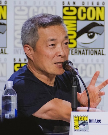 Hombre asiático de mediana edad con cabello gris hablando en un micrófono. Se ven una botella de agua, un cartel con el logo de Comic-Con y el nombre Jim Lee