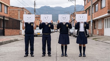 Cuatro niños con uniforme escolar cubren sus caras con carteles que muestran caras dibujadas: triste, confusa, enojada y feliz, en una calle urbana.
