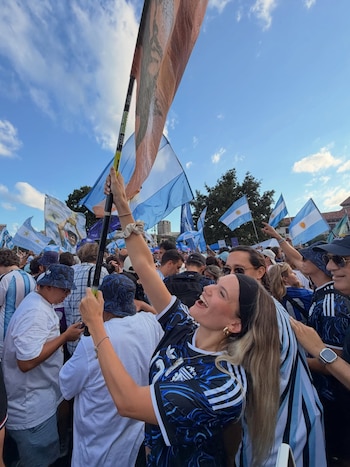 Multitud de personas con camisetas y banderas de Argentina. Una mujer rubia sonríe y levanta una bandera argentina en primer plano
