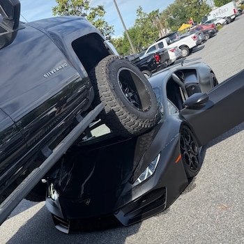 Camioneta Chevrolet Silverado negra montada sobre un Lamborghini Huracán negro en un estacionamiento. El Lamborghini tiene la puerta del conductor abierta