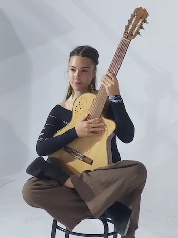 Una persona con cabello largo toca una guitarra acústica con los ojos cerrados frente a un micrófono en un escenario iluminado