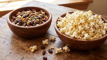 Dos cuencos de madera llenos de frutos secos variados y palomitas de maíz están sobre una mesa de madera, con algunos granos de maíz y pasas esparcidos.