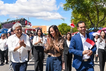 María René Soruco, de cabello oscuro, aplaude sonriente, vistiendo una blusa satinada marrón y pantalón azul, rodeada de gente en un evento al aire libre