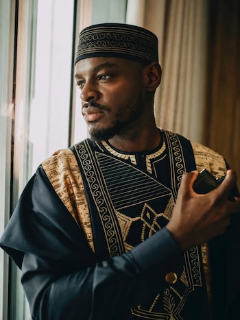 Hombre con barba y gorro kufi, vestido con una túnica negra y dorada bordada, mira por una ventana, con un objeto oscuro en su mano derecha