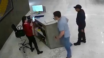Mujer con teléfono, dos hombres, mostrador, monitor de computadora, silla de oficina en espacio interior con piso claro.