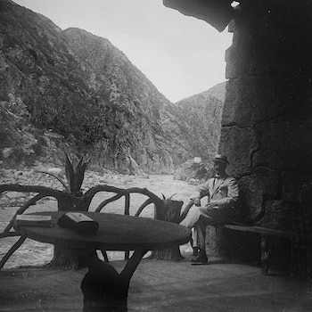 Fotografía en blanco y negro de un hombre sentado junto a una mesa en un mirador con barandilla rústica frente a un río entre montañas