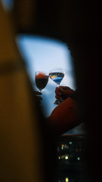 Dos personas, una con Amaretto Sour y otra con Spritz, levantan sus copas en un brindis, con un fondo borroso de cielo azul y luces de la ciudad