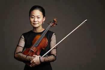 Retrato de una mujer asiática sonriendo, sosteniendo un violín de madera rojiza y un arco en sus brazos, vestida con una blusa negra de manga larga. El fondo es liso y oscuro