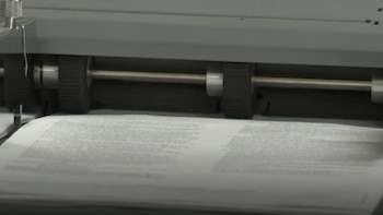 Primer plano de una máquina gris con rodillos negros que alimentan una hoja de papel blanco impresa con texto, visible en proceso