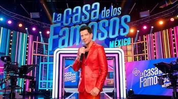 Cantante Yahir de pie, sonriendo y sosteniendo un micrófono, viste traje rojo de cuero en un escenario con el logo "La Casa de los Famosos México" y luces coloridas.