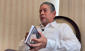 Un hombre de cabello gris y barba, con camisa blanca, está sentado sosteniendo un libro cuya portada muestra un rostro, con un micrófono sujeto a su ropa