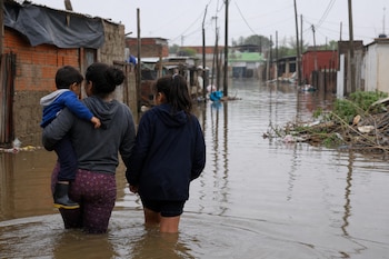 Una mujer con un niño en brazos y una niña caminan de espaldas por una calle inundada entre viviendas precarias y postes de luz.