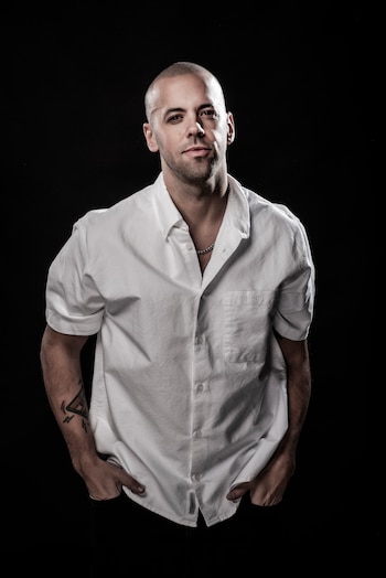 Hombre calvo con barba, camisa blanca de manga corta, cadena plateada, tatuaje en brazo y manos en los bolsillos, sobre fondo negro