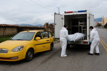 Dos personas con trajes blancos de protección introducen una camilla con un cuerpo cubierto en un furgón blanco junto a un taxi amarillo.