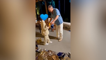 Un hombre de camisa a rayas entrega un objeto a un niño vestido con traje beige junto a globos y adornos en el suelo