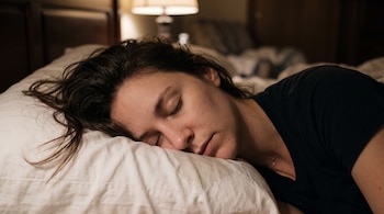 Plano cercano del rostro de una persona dormida sobre una almohada blanca con gotas de sudor en la piel y una lámpara al fondo.