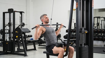Hombre con camiseta gris y pantalón negro, sentado en máquina de lat pulldown, sujeta la barra, en gimnasio con equipamiento y espejos.