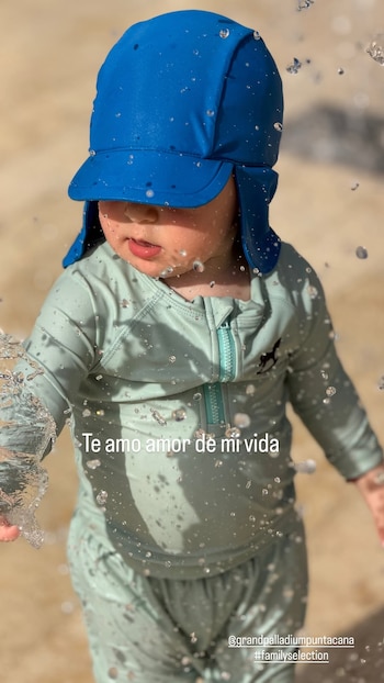Un bebé con gorro azul y traje de baño de manga larga juega entre chorros de agua con el mensaje Te amo amor de mi vida