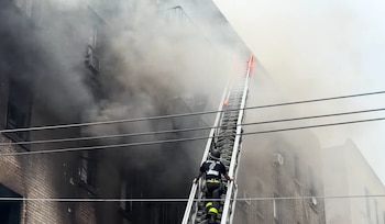 Un bombero con casco y uniforme asciende por una escalera extensible apoyada en un edificio de ladrillo del que sale humo denso y gris