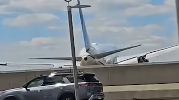 Vista trasera de un avión de pasajeros blanco con cola azul, alas extendidas, detrás de una barrera gris y un SUV gris en primer plano, bajo cielo con nubes
