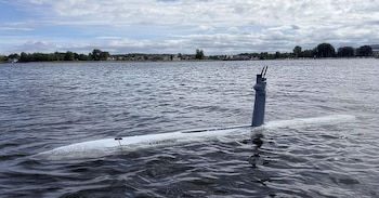 Vehículo sumergible blanco y gris emergiendo parcialmente del agua, con un mástil que tiene dos antenas. Fondo de mar, orilla con árboles y casas bajo cielo nublado