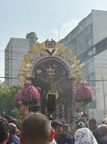 El anda procesional del Señor de los Milagros adornada con flores, rodeada por personas frente a edificios urbanos