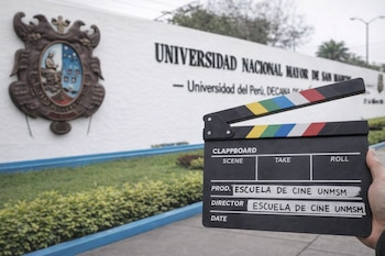 Una mano sostiene un clapperboard negro con franjas de colores frente al escudo y el nombre Universidad Nacional Mayor de San Marcos en una pared blanca.