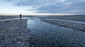 Un pescador solitario de espaldas, con caña, en la orilla izquierda de un río trenzado. Múltiples canales de agua transparente y bancos de grava bajo un cielo nublado al atardecer.