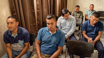 Seis hombres sentados en sillas negras con cortinas marrones al fondo. Uno de los hombres lleva esposas en las muñecas y sujeta una botella transparente.