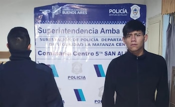 Un hombre joven de cabello oscuro y chaqueta negra posa frente a un banner azul y blanco. A la izquierda, la espalda de un hombre con uniforme policial