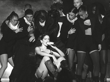 Una mujer con un micrófono sentada, rodeada por diez personas de pie, en un escenario oscuro. Es una foto en blanco y negro