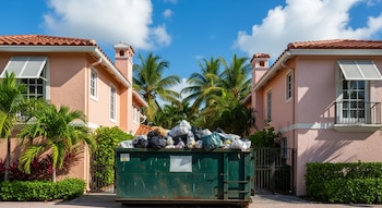 Un contenedor de basura verde lleno de bolsas de residuos se ubica frente a dos casas rosadas con tejados de teja, rodeadas de palmeras y vegetación.