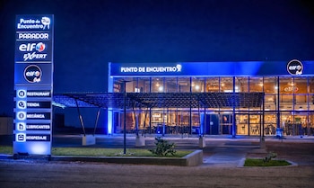 Edificio comercial iluminado de noche. Un cartel vertical muestra los servicios de Punto de Encuentro: restaurante, tienda, mecánica, lubricantes y hospedaje