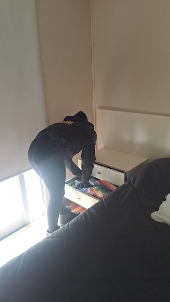 Persona con uniforme oscuro buscando en un cajón de un mueble blanco con tiradores plateados. El cajón contiene ropa y un objeto de color arcoíris. A la izquierda hay una ventana con luz brillante