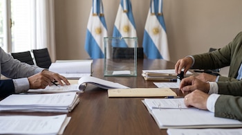 Primer plano de manos sobre una mesa de madera con documentos, papeles, plumas y una urna de vidrio. Al fondo se observan banderas de Argentina.