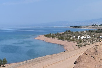 Vista panorámica de una playa de arena junto a un vasto cuerpo de agua azul. Hay vegetación, tiendas de campaña blancas y una gran duna de arena en primer plano