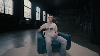 Un hombre sonriente sentado en un sillón azul, sosteniendo una claqueta de cine. El fondo es un estudio grande y oscuro con ventanales enrejados