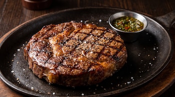 Ojo de bife asado condimentado con pimienta y sal gruesa en una sartén de hierro, junto a un recipiente con chimichurri.