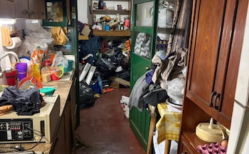 Habitación con armarios, estanterías, encimera de cocina, fregadero, puerta abierta, suelo de baldosas, ropa, bolsas de basura, cajas, botellas y objetos diversos