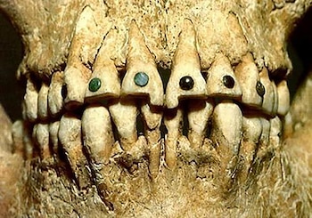 Primer plano de dientes humanos antiguos, con incrustaciones de pequeñas gemas redondas verdes, azules y negras visibles en la parte frontal de varias piezas dentales superiores
