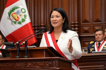 Mujer con blazer blanco y banda bicolor habla ante dos micrófonos sobre un podio de madera, con una bandera de Perú detrás y un hombre en segundo plano.