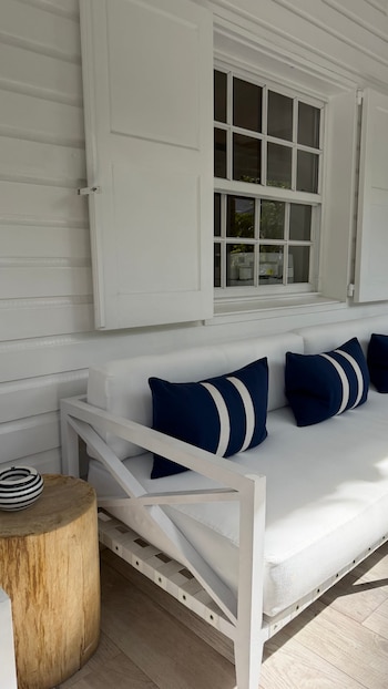 Un sofá blanco con cojines azul marino de rayas blancas sobre un suelo de madera, junto a una mesa de tronco y una ventana con persianas abiertas